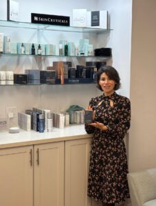 Woman in black floral dress showcasing skincare product in front of shelves of products
