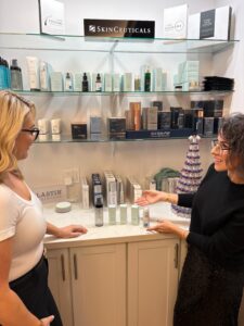 Brunette woman with glasses in black dress showing skincare products to blond woman in glasses and white t-shirt