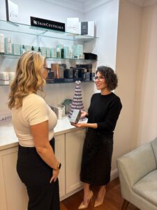 Brunette woman with glasses in black dress holding out skincare product to blond woman in glasses and white t-shirt